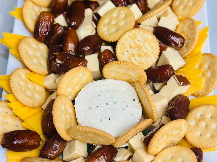 A platter with round crackers, yellow and white cheese slices, dates, and a round wheel of herbed cheese in the center, arranged in a decorative pattern.