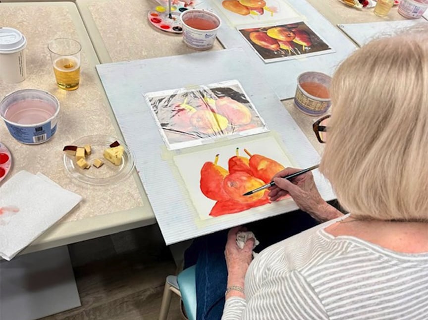 A woman with blonde hair and glasses paints a watercolor image of red apples at a table, surrounded by art supplies, reference photos, drinks, and a plate with apple slices and cheese.