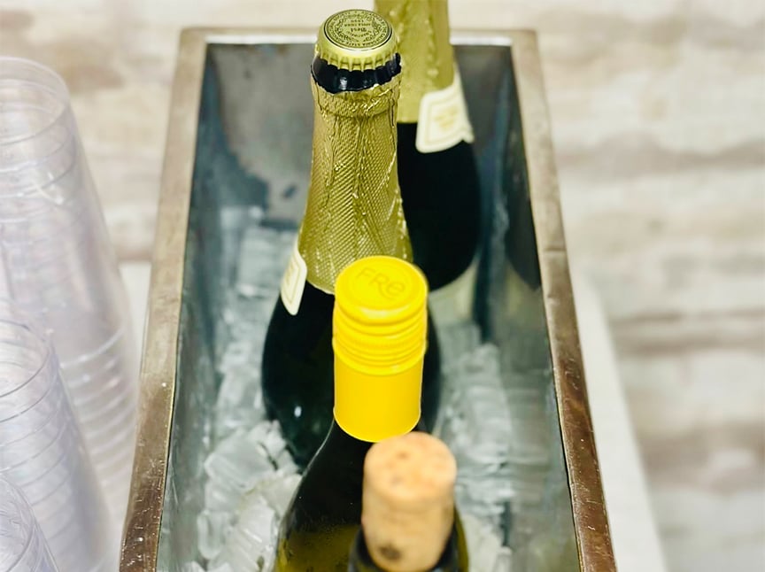 Three bottles of wine or champagne in an ice-filled metal bucket, with plastic cups stacked nearby. The bottles have different caps: a cork, a yellow screw cap, and a mesh-wrapped top.