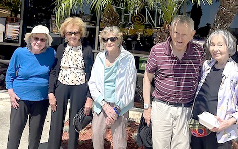 Five older adults, three women and two men, stand together outdoors smiling in front of a storefront with palm trees and sunshine. They are dressed in casual clothing and sunglasses, enjoying a bright day.
