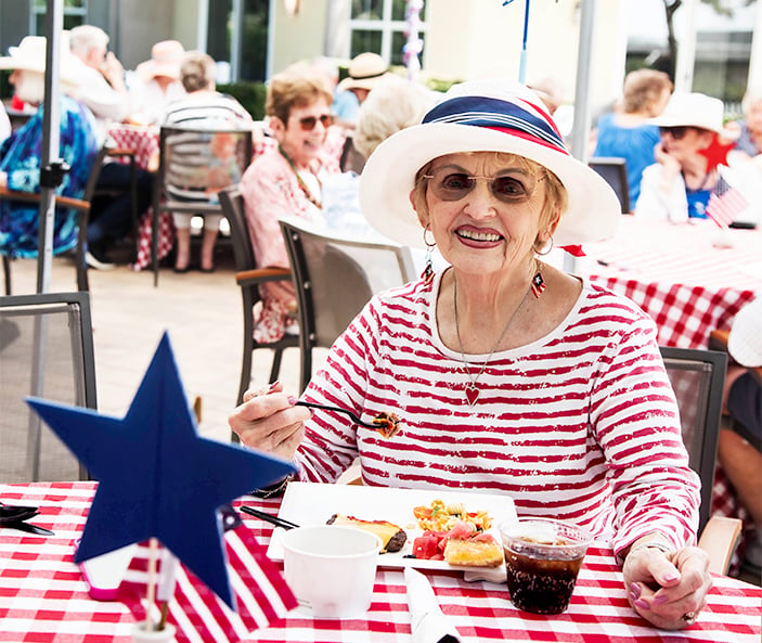 An older woman wearing a white hat with red and blue stripes smiles while eating at an outdoor picnic. She is seated at a table with a red checkered tablecloth and patriotic decorations. Other people are in the background.