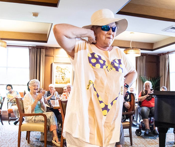 An older woman wearing a sunhat, sunglasses, and a T-shirt printed with a bikini design strikes a playful pose in front of a smiling, clapping audience in a bright, cheerful room.