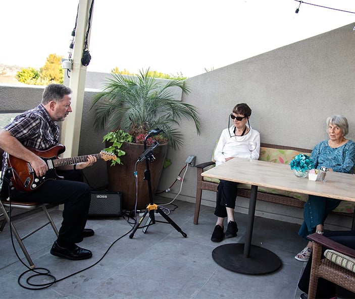 A man plays an electric guitar for two seated women on a patio. The women watch and listen, one wearing sunglasses. There are potted plants, a table with flowers, and a wall in the background.