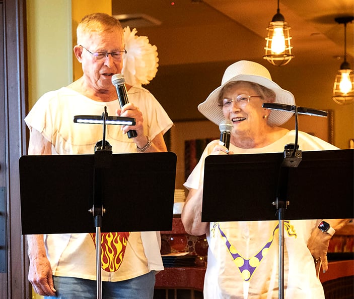 An older man and woman sing into microphones, standing side by side. Both wear novelty t-shirts with swimsuit prints; the woman also wears a white sunhat. They look happy and are standing behind black music stands indoors.