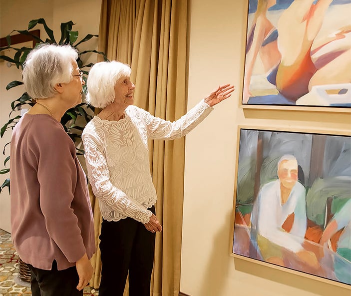 Two older women stand indoors, one pointing toward colorful abstract paintings displayed on the wall. Both appear engaged and smiling, with plants and curtains in the background.