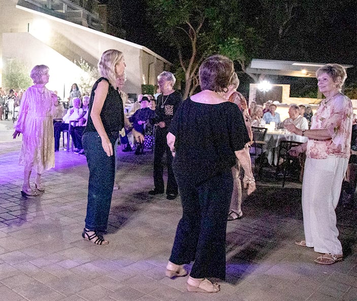 A group of older women dance together outdoors at night, surrounded by seated people watching. The area is lit with purple and white lights, and trees and buildings are visible in the background.