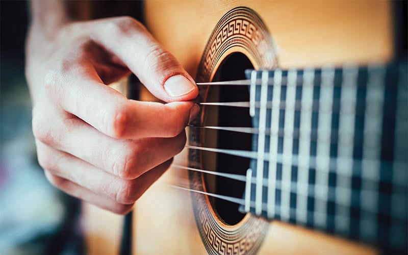 A close-up of a hand plucking the strings of an acoustic guitar, focusing on the fingers and the guitars sound hole with decorative patterns around it.