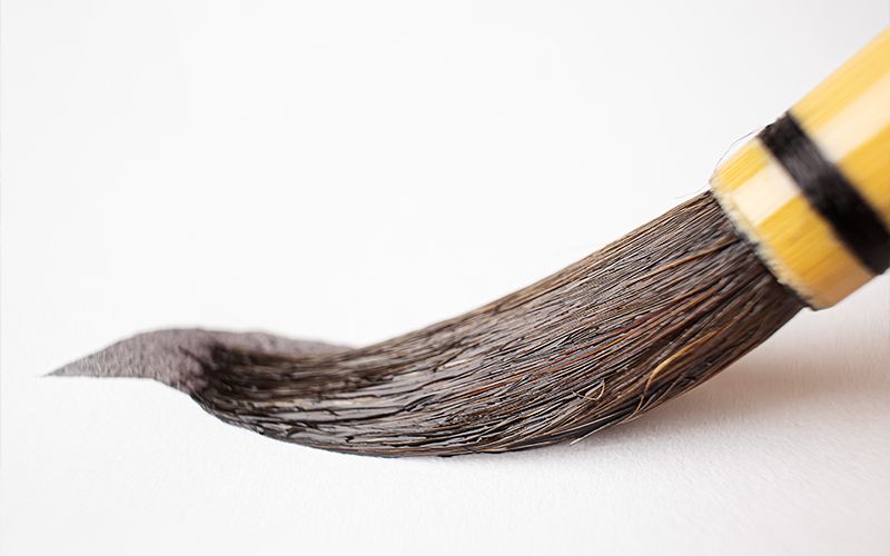 A close-up of a calligraphy brush with black bristles, sweeping black ink onto white paper. The handle is yellow with black bands near the bristles.