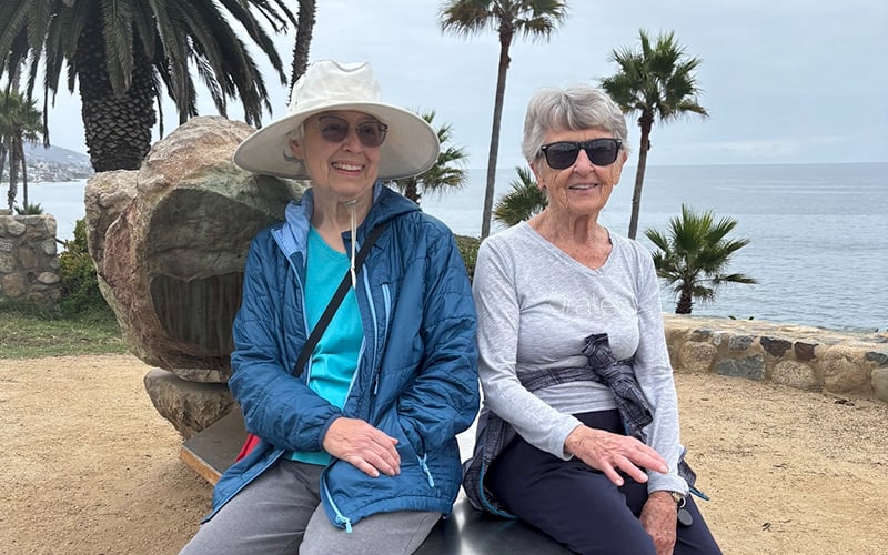 Two older women wearing sunglasses and casual clothes sit on a bench by the ocean, palm trees and a large rock behind them, on a cloudy day.