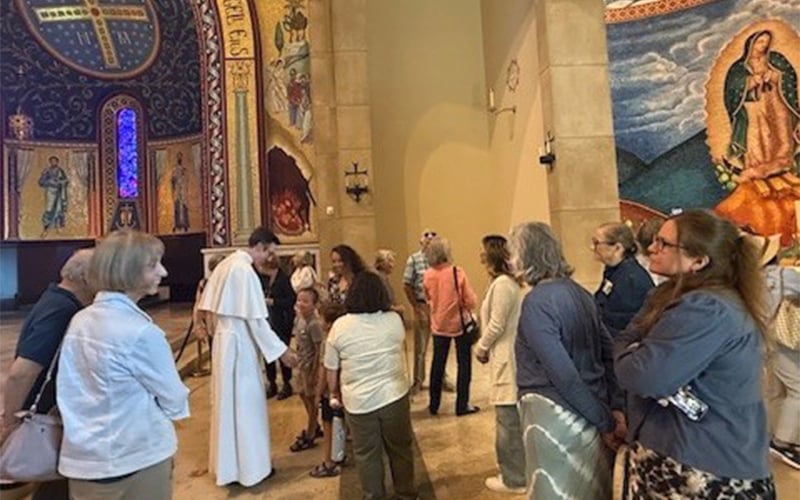 A group of people, including a priest in white robes, gather and converse inside a church decorated with colorful murals and religious art.