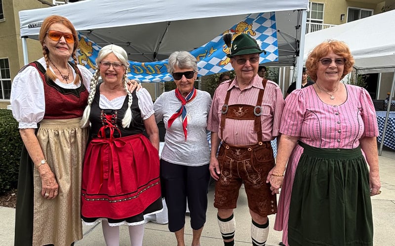 Five older adults stand together outside under a canopy, three women wearing traditional German dresses, one man in lederhosen and a hat, and another woman in casual clothes, smiling for the camera.
