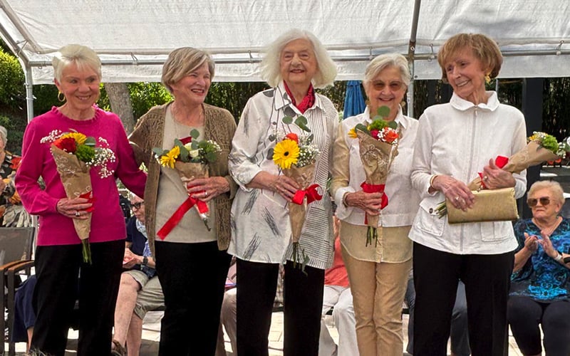 Five smiling elderly women stand side by side outdoors, each holding a bouquet of flowers. They are dressed in light, casual clothing, and people are seated and watching them in the background under a canopy.
