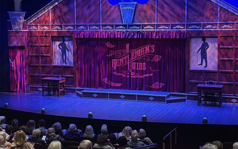 A theater stage set for A Gentlemans Guide to Love & Murder features red curtains, two framed silhouettes on either side, small tables and chairs on stage, and an audience waiting for the show to begin.
