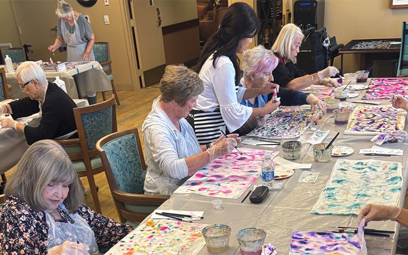 A group of older adults sit around tables, painting colorful patterns on fabric. A younger woman assists one participant. The room is bright and filled with art supplies and finished pieces.