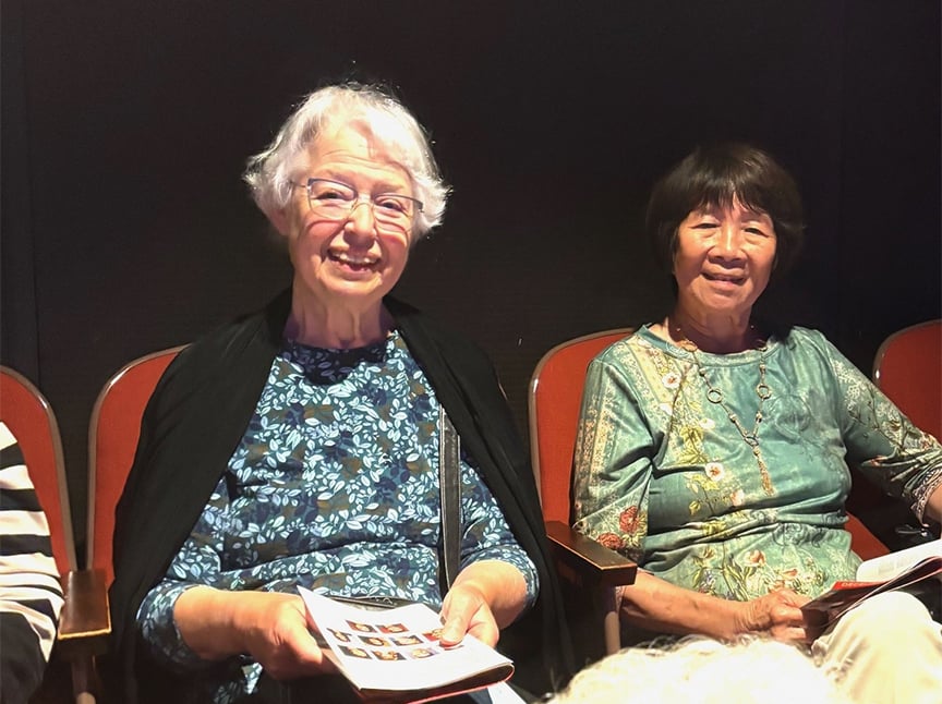 Two older women sit side by side in red theater seats, smiling at the camera. Both are holding programs. The woman on the left has short white hair and glasses; the woman on the right has short dark hair.