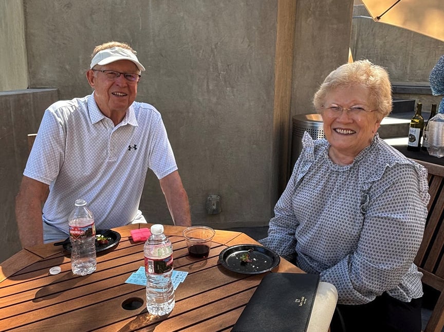 An older man and woman sit at a wooden outdoor table, smiling. There are water bottles, plates with food remnants, drinks, and notes on the table. They are casually dressed and appear to be enjoying a sunny day.