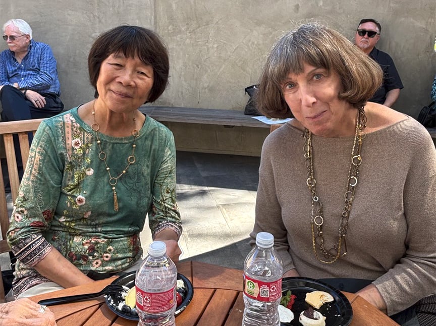 Two women sit at an outdoor table with plates of food and water bottles, smiling at the camera. A man sits on a bench in the background against a concrete wall. Sunlight casts shadows across the scene.