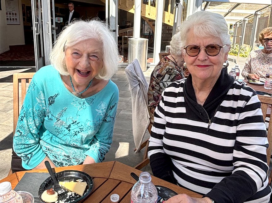 Two older women sit at an outdoor table, smiling. One wears a turquoise top, the other wears a black-and-white striped sweater. Plates of dessert and bottled water are on the table. Sunlight brightens the scene.