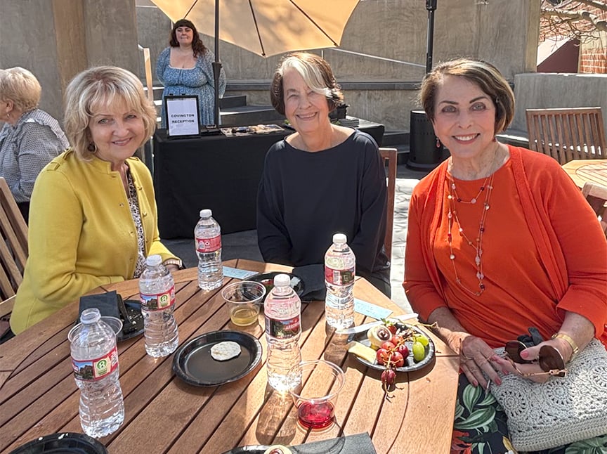 Three smiling women sit at a round wooden outdoor table with snacks, fruit, and bottled water. Another woman stands in the background near a sign and umbrellas shade the sunny area.