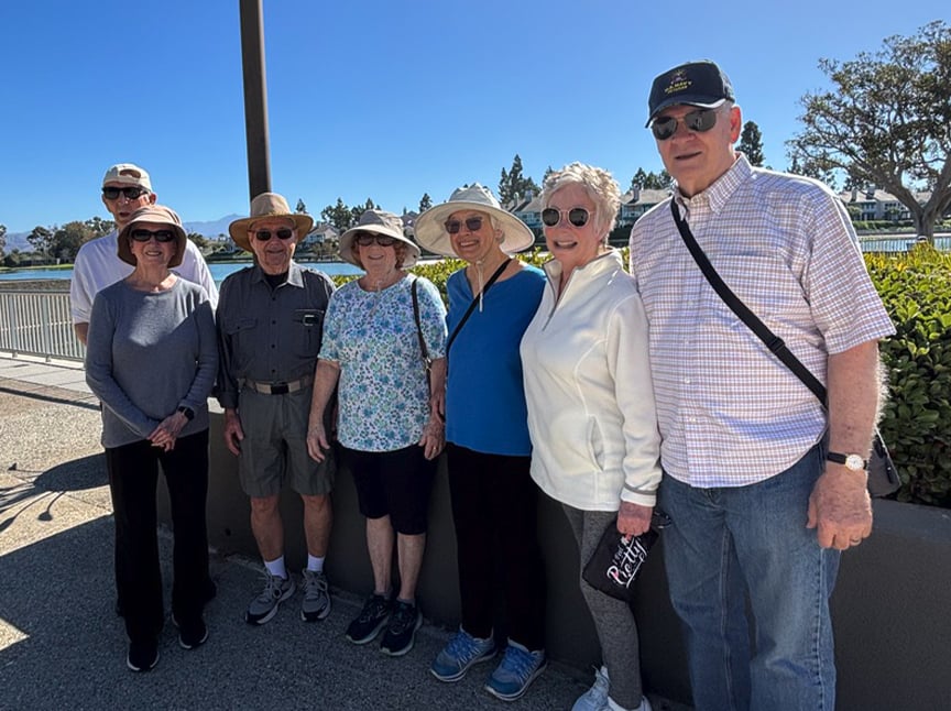 A group of eight older adults, wearing hats and sunglasses, stand together outdoors on a sunny day, smiling at the camera. Trees, bushes, and a blue sky are visible in the background.