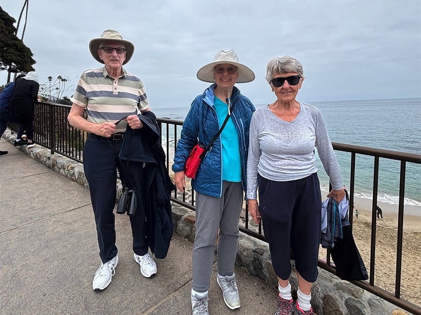 Three older adults stand and smile on a seaside walkway near a metal railing, with the ocean and sandy beach in the background. They wear casual hats, jackets, and sunglasses on a cloudy day.