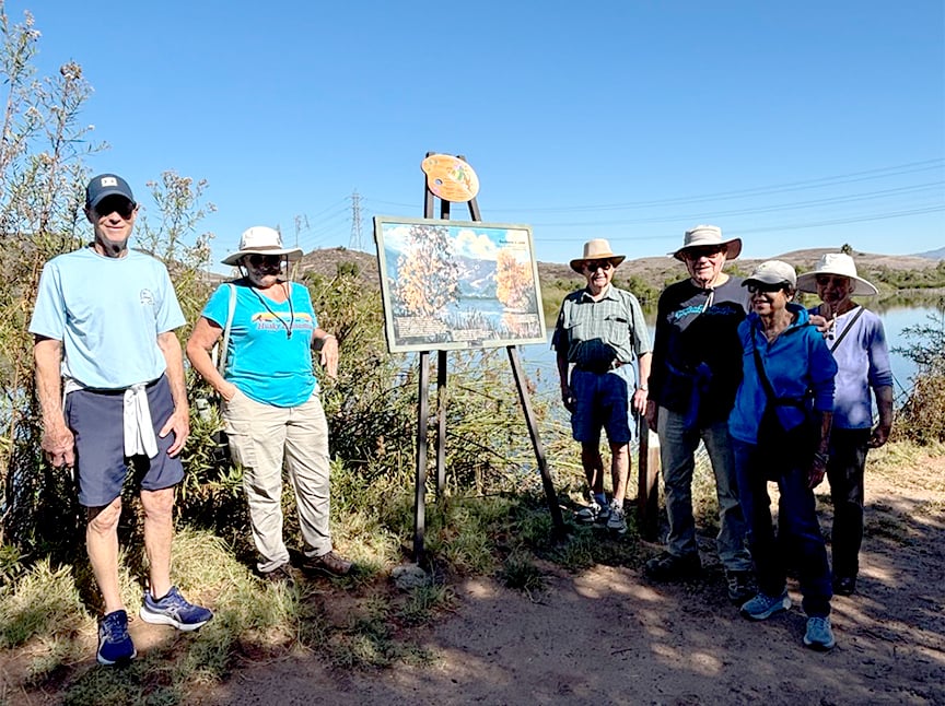 A group of six people in casual outdoor clothing and hats stand on a sunny trail beside a sign with a nature scene, surrounded by bushes and a body of water under a clear blue sky.