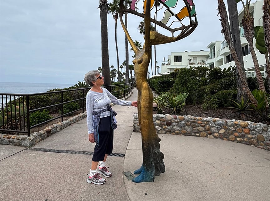 An older woman stands on a paved path by the ocean, touching the hand of a colorful, abstract female sculpture. Palm trees, modern white buildings, and lush greenery are in the background.