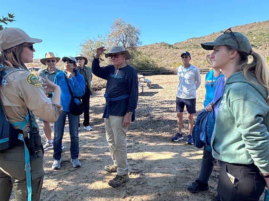 A group of people, including a park ranger, stand outdoors on a sunny day having a conversation. Some wear hats and sunglasses. Trees and hills are in the background.