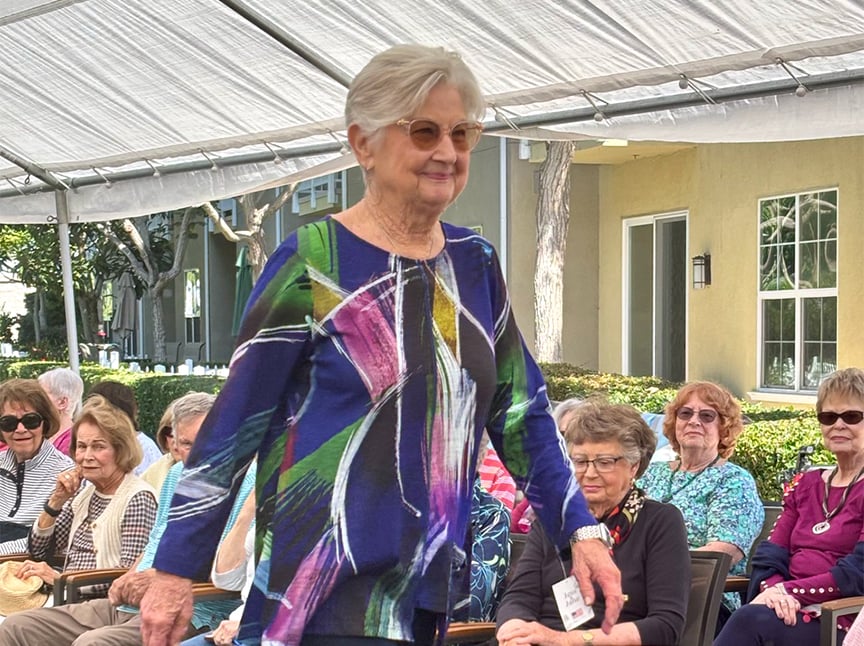An older woman wearing sunglasses and a colorful long-sleeve blouse walks outdoors under a canopy, with a group of seated older women watching and smiling in the background.
