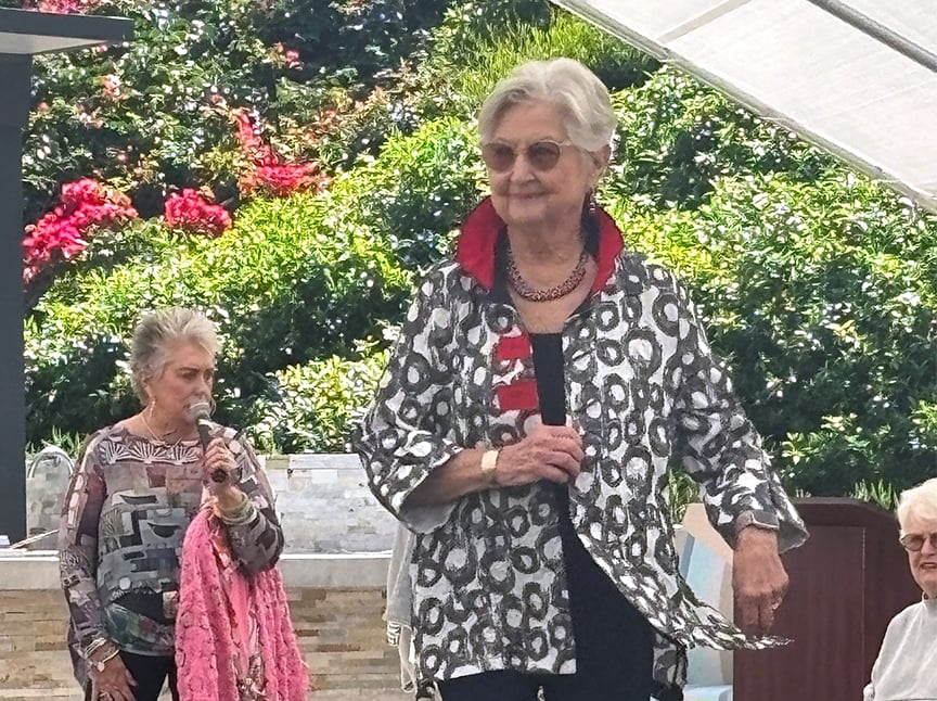 An older woman wearing a patterned jacket walks outdoors, smiling, with another woman speaking into a microphone nearby. Bright greenery and flowers are in the background, and other people sit nearby.