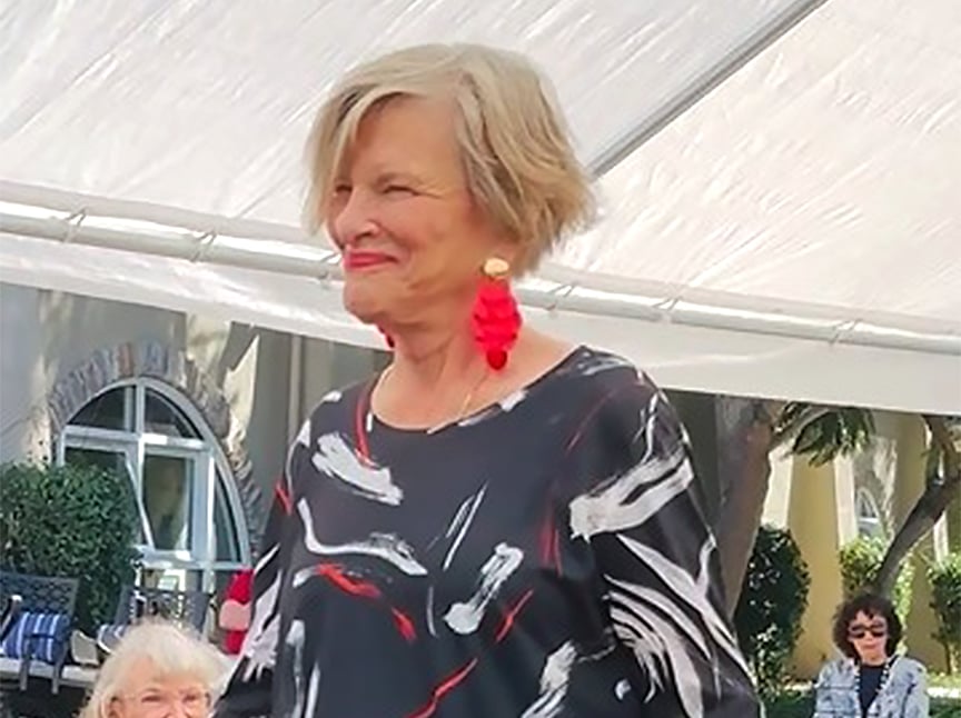 An older woman with short, light hair smiles while wearing bold red earrings and a black patterned top. She stands outdoors under a white canopy, with other people seated in the background.