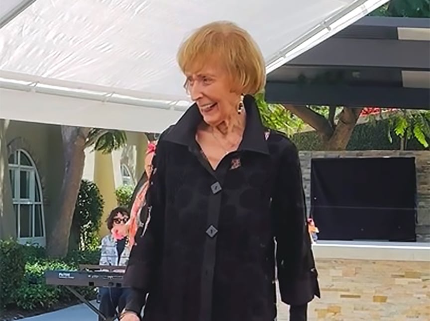 An elderly woman with short, light hair and a black jacket smiles outdoors under a white canopy, with trees and a person playing keyboard in the background.