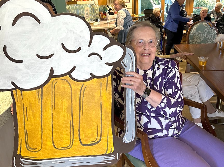 An elderly woman in a purple and white outfit smiles while holding a large, cartoon-style cutout of a beer mug with foam. She is seated in a lively room with other people in the background.