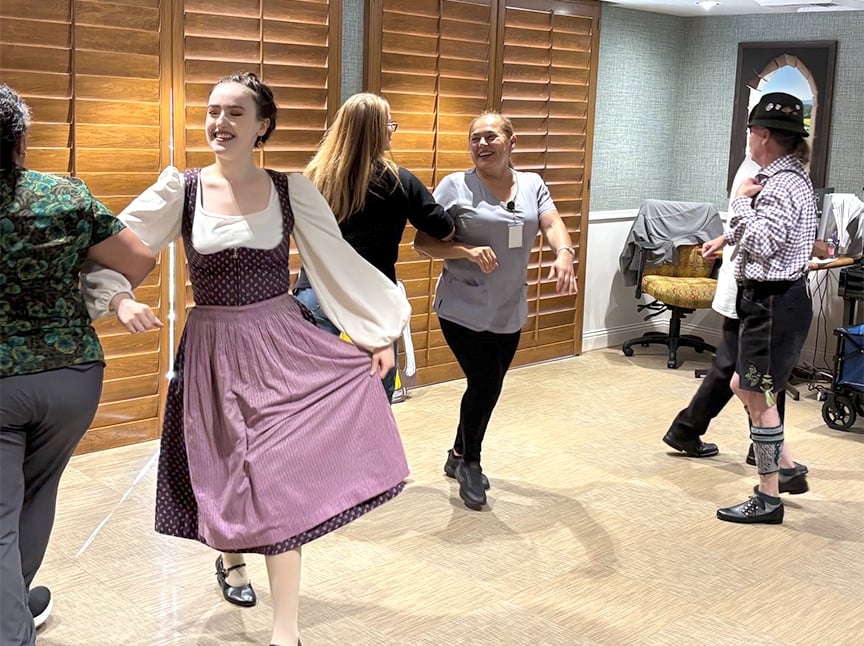 A group of people dance indoors, with one woman in the foreground wearing a traditional dress. Others, casually dressed, join in the dance. Wood-paneled shutters and chairs are visible in the background.