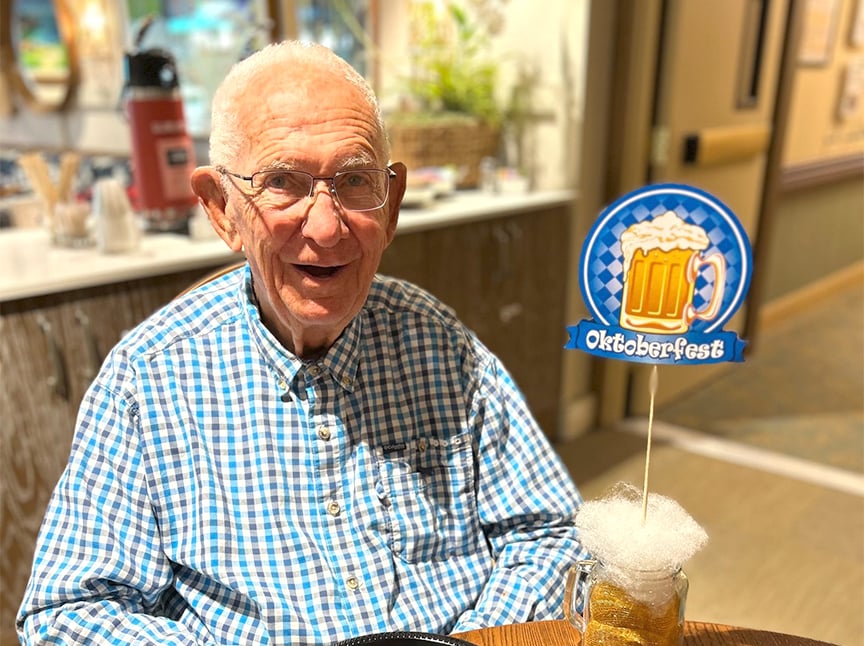 An elderly man in glasses and a blue checkered shirt sits at a table, smiling. In front of him is a decorative Oktoberfest sign with a beer mug image on it. The background shows a bright indoor setting.