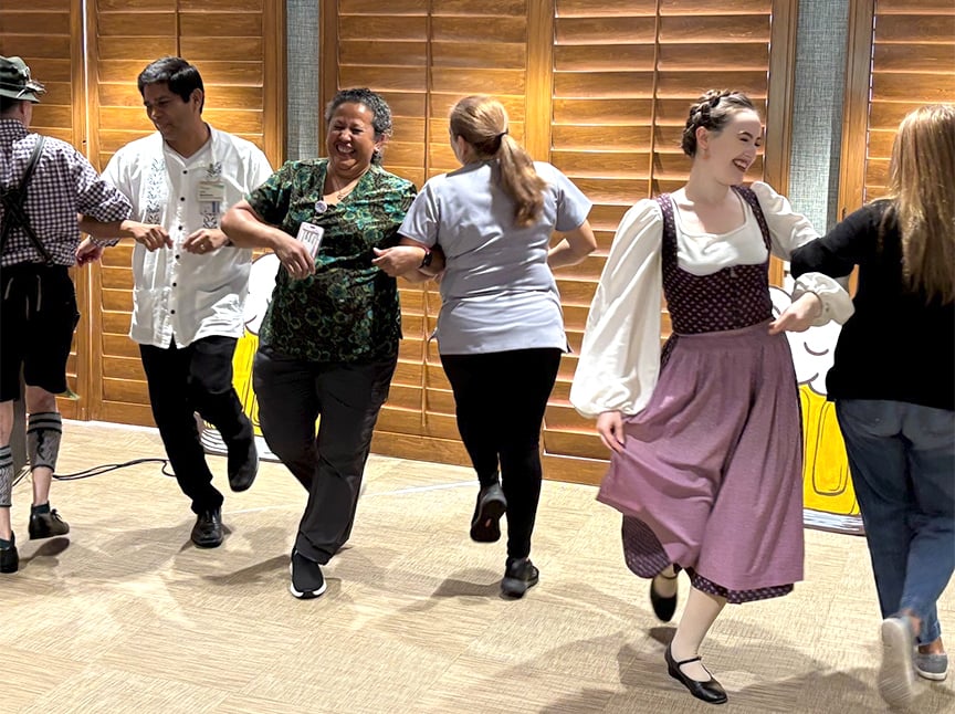A group of people, some in traditional German attire, dance in pairs indoors on a wood floor, holding hands and smiling, with wooden shutters in the background.