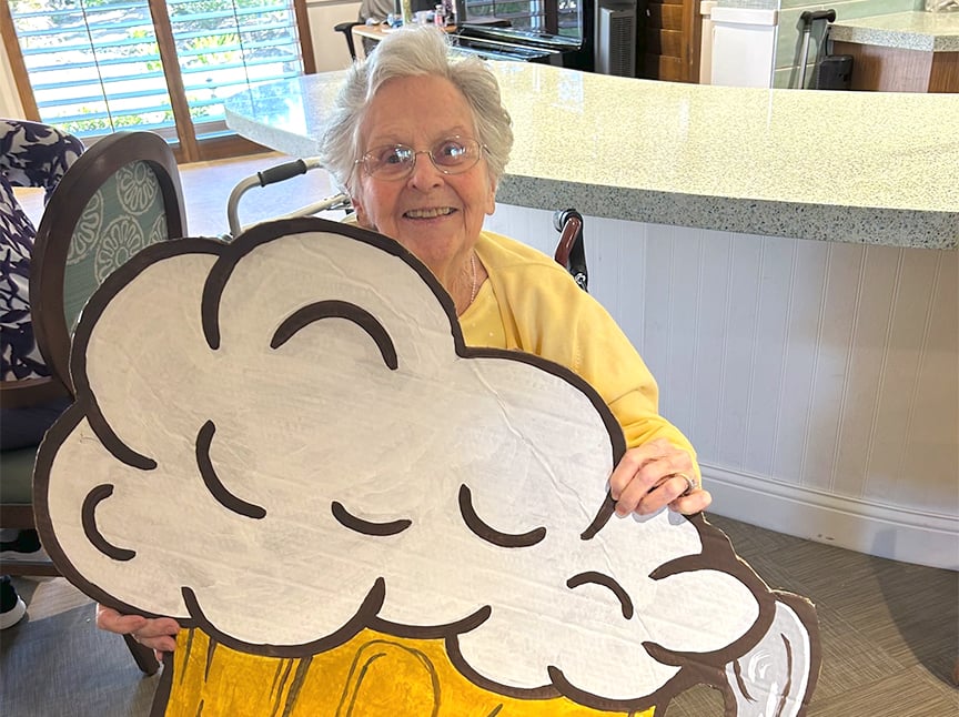 An elderly woman smiling while holding a large cardboard cutout of a beer mug with frothy foam, sitting in a bright, modern kitchen.