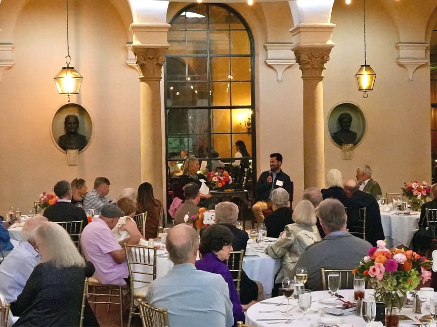 A man speaks into a microphone to an audience seated at round tables in an elegant hall with arched columns, floral centerpieces, and warm lighting. Bust sculptures are mounted on the walls.