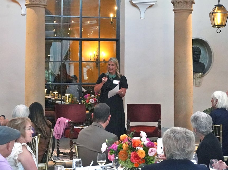 A woman speaks into a microphone while standing on a stage in front of large windows at a formal indoor event. Attendees are seated at tables with floral centerpieces, listening to her. The atmosphere is elegant and festive.