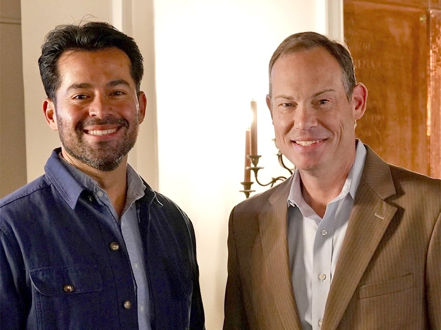 Two men smiling and standing side by side indoors, one in a blue shirt and the other in a brown suit jacket, with a lit wall sconce visible in the background.