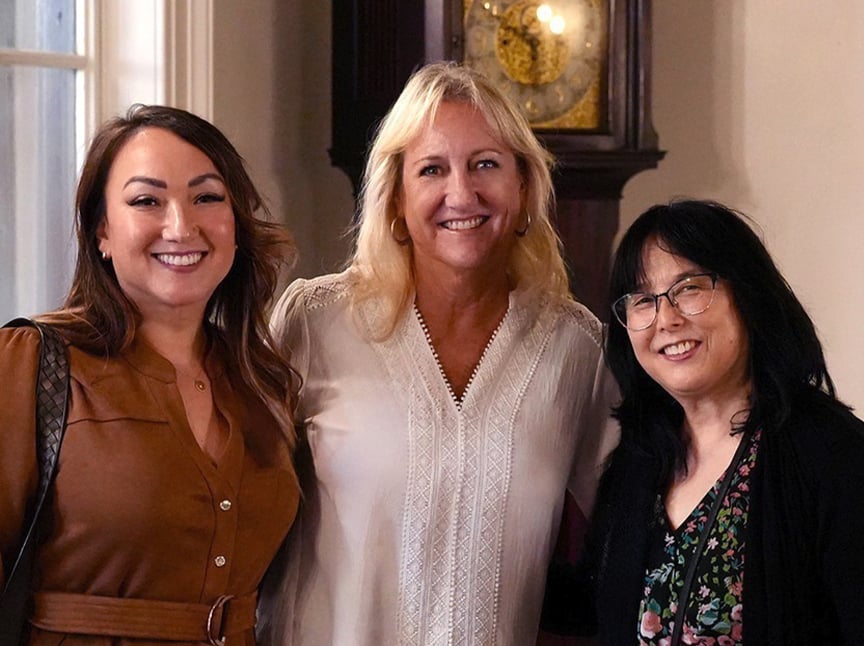 Three women stand close together indoors, smiling at the camera. The woman on the left wears a brown dress, the center woman wears a light embroidered blouse, and the woman on the right has glasses and a floral top. A clock is visible behind them.