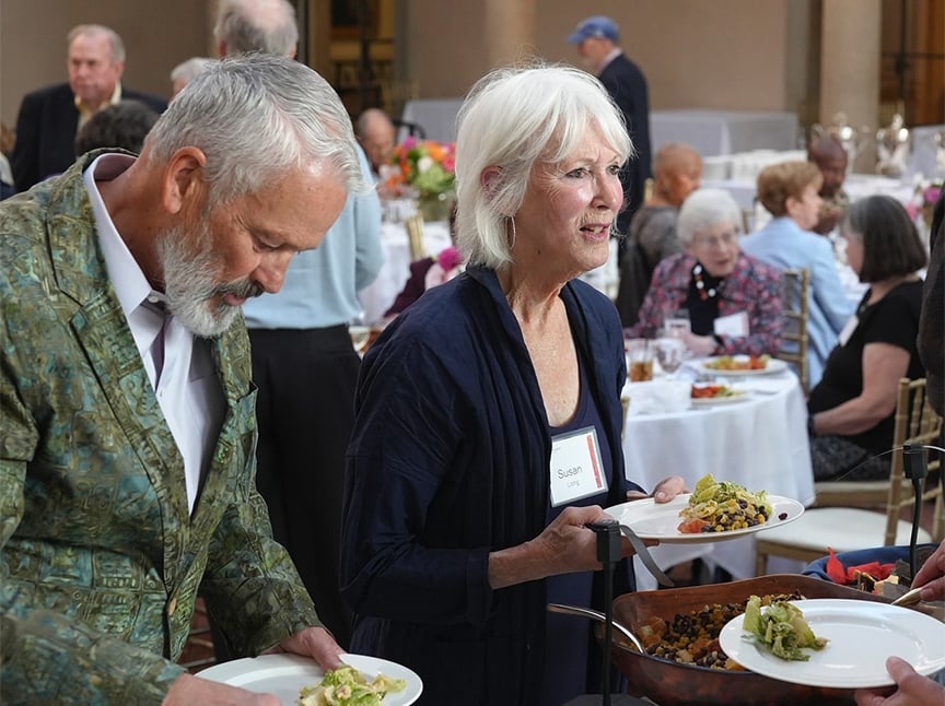 Two older adults, a man in a patterned jacket and a woman with white hair wearing a name tag, are serving themselves food from a buffet at a formal indoor event with other guests seated at round tables in the background.