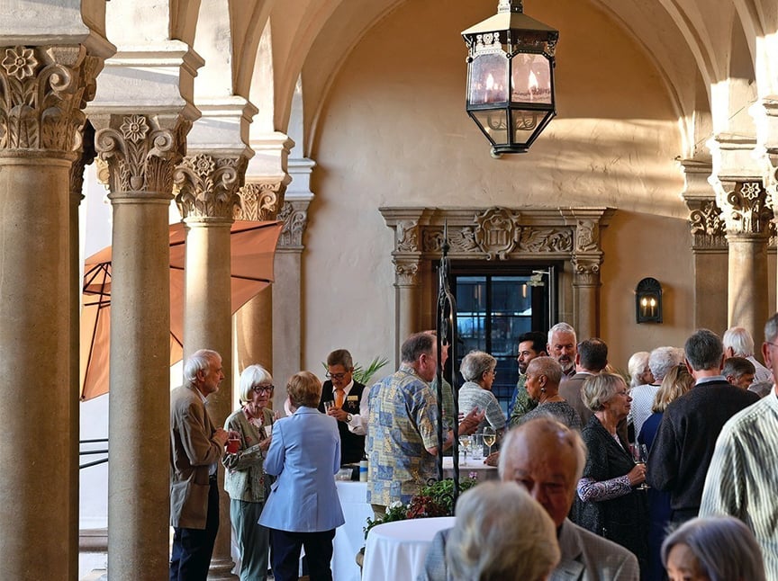 A group of people socialize and mingle at a formal indoor gathering, surrounded by ornate columns and arches with warm lighting. Tables with white tablecloths and an orange umbrella are visible in the background.