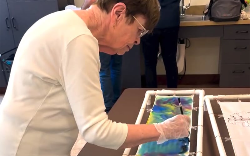 An older woman wearing glasses and gloves paints a colorful design on fabric stretched over a frame in an art classroom. Other people and supplies are visible in the background.