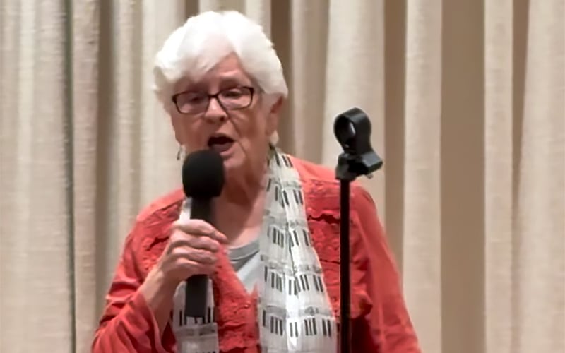 An elderly woman with short white hair, glasses, and a piano key patterned scarf holds a microphone while speaking or singing in front of beige curtains. A microphone stand is beside her.