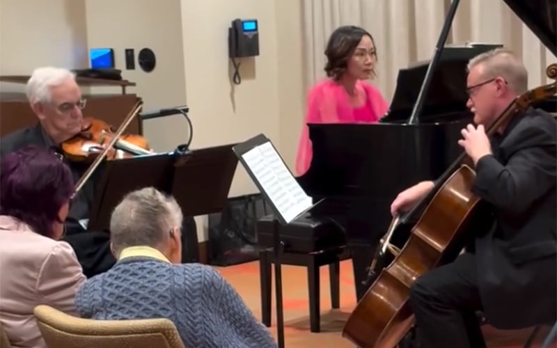 Three musicians perform indoors: a violinist, a pianist, and a cellist. Two audience members sit in the foreground, listening attentively. The musicians are focused on their sheet music and instruments.