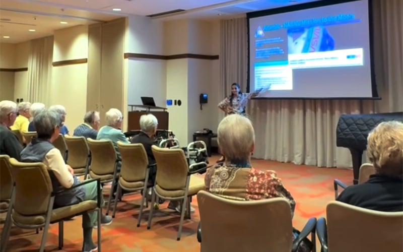 A woman gives a presentation to a seated audience of older adults in a conference room, gesturing toward a projection screen with informational slides.