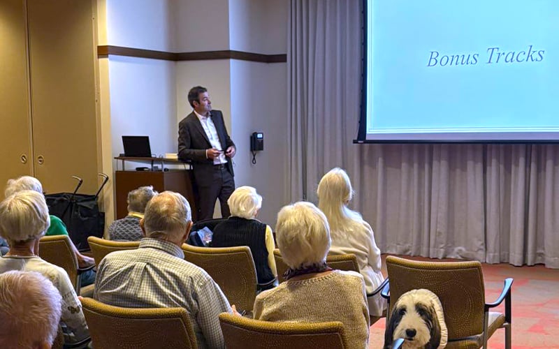A man in a suit presents a slideshow titled Bonus Tracks to an audience of older adults seated in chairs, with some grey and white-haired attendees visible from the back.