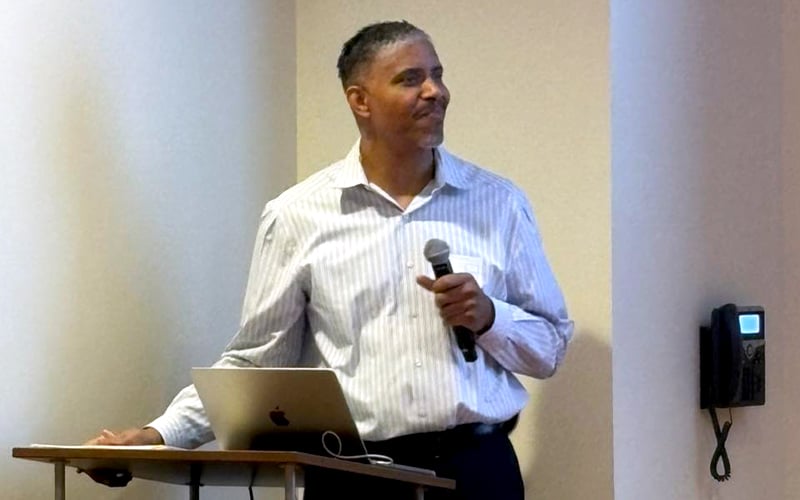 A man in a striped shirt stands at a podium with a laptop, holding a microphone and smiling, as if speaking or presenting. A phone is mounted on the wall behind him.
