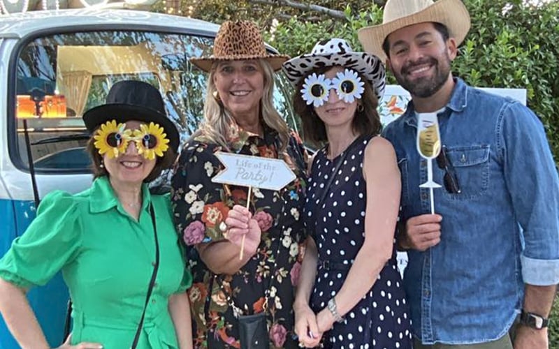 Four people dressed in playful, colorful outfits and hats pose together outdoors. Two hold props: a Life of the Party! sign and a paper wine glass. A blue van is in the background. All are smiling and festive.